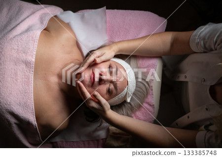 Close up of a mature woman receiving a relaxing facial massage from a professional female therapist in a modern spa. The client lies on a treatment bed with a towel and headband, enjoying skincare Close up of a mature woman receiving a relaxing facial massage from a professional female therapist in a modern spa. The client lies on a treatment bed with a towel and headband, enjoying skincare 133385748