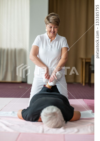 Professional female physiotherapist helping an older woman stretch her legs while lying face down on a mat. Rehabilitation and physiotherapy concept focusing on mobility recovery, flexibility, and 133385755