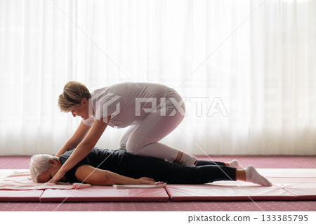 Professional female physiotherapist supporting an older woman during a therapeutic stretching exercise on a mat. Rehabilitation and physiotherapy concept focused on mobility, flexibility, recovery Professional female physiotherapist supporting an older woman during a therapeutic stretching exercise on a mat. Rehabilitation and physiotherapy concept focused on mobility, flexibility, recovery 133385795