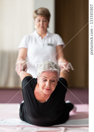 Professional female physiotherapist supporting an older woman during a therapeutic stretching exercise on a mat. Rehabilitation and physiotherapy concept focused on mobility, flexibility, recovery Professional female physiotherapist supporting an older woman during a therapeutic stretching exercise on a mat. Rehabilitation and physiotherapy concept focused on mobility, flexibility, recovery 133385807
