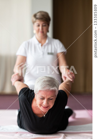 Professional female physiotherapist supporting an older woman during a therapeutic stretching exercise on a mat. Rehabilitation and physiotherapy concept focused on mobility, flexibility, recovery Professional female physiotherapist supporting an older woman during a therapeutic stretching exercise on a mat. Rehabilitation and physiotherapy concept focused on mobility, flexibility, recovery 133385809