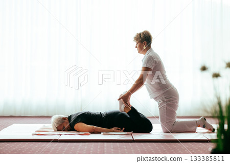Professional female physiotherapist helping an older woman stretch her legs while lying face down on a mat. Rehabilitation and physiotherapy concept focusing on mobility recovery, flexibility, and Professional female physiotherapist helping an older woman stretch her legs while lying face down on a mat. Rehabilitation and physiotherapy concept focusing on mobility recovery, flexibility, and 133385811