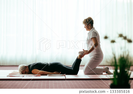 Professional female physiotherapist helping an older woman stretch her legs while lying face down on a mat. Rehabilitation and physiotherapy concept focusing on mobility recovery, flexibility, and 133385816