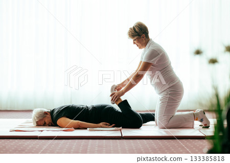Professional female physiotherapist helping an older woman stretch her legs while lying face down on a mat. Rehabilitation and physiotherapy concept focusing on mobility recovery, flexibility, and 133385818