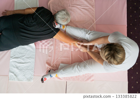 A physiotherapist assisting an older woman with a shoulder and arm stretch while lying on a mat. Rehabilitation and physiotherapy session concept focusing on mobility, flexibility, pain relief, and 133385822