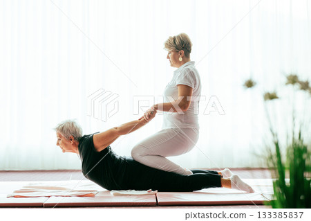 Professional female physiotherapist supporting an older woman during a therapeutic stretching exercise on a mat. Rehabilitation and physiotherapy concept focused on mobility, flexibility, recovery Professional female physiotherapist supporting an older woman during a therapeutic stretching exercise on a mat. Rehabilitation and physiotherapy concept focused on mobility, flexibility, recovery 133385837