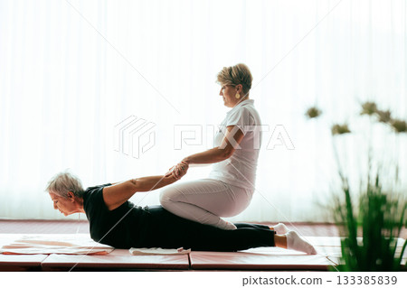Professional female physiotherapist supporting an older woman during a therapeutic stretching exercise on a mat. Rehabilitation and physiotherapy concept focused on mobility, flexibility, recovery 133385839