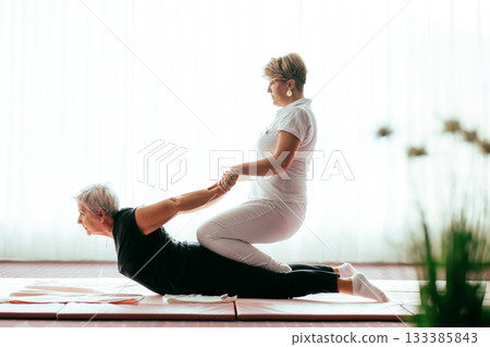 Professional female physiotherapist supporting an older woman during a therapeutic stretching exercise on a mat. Rehabilitation and physiotherapy concept focused on mobility, flexibility, recovery 133385843