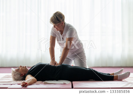 Professional female physiotherapist assisting an older woman lying on a mat during a therapeutic rehabilitation session. Wellness and physiotherapy concept highlighting recovery, mobility, pain relief 133385856