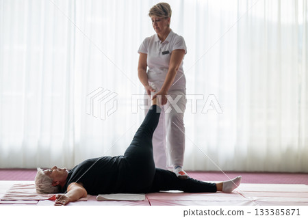 Professional female physiotherapist assisting an older woman with a leg stretch while lying on a mat during a rehabilitation session. Concept of physiotherapy, mobility recovery, flexibility, and 133385871