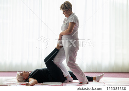 Professional female physiotherapist assisting an older woman with a leg stretch while lying on a mat during a rehabilitation session. Concept of physiotherapy, mobility recovery, flexibility, and 133385888