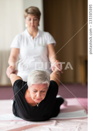 Professional female physiotherapist supporting an older woman during a therapeutic stretching exercise on a mat. Rehabilitation and physiotherapy concept focused on mobility, flexibility, recovery 133385895