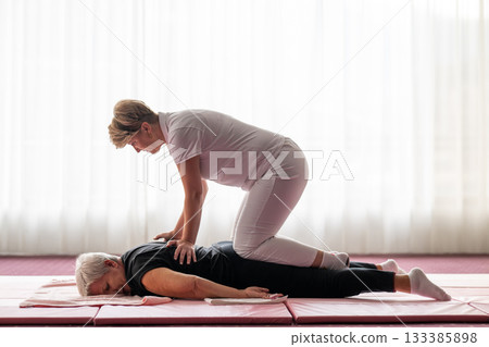Professional female physiotherapist supporting an older woman during a therapeutic stretching exercise on a mat. Rehabilitation and physiotherapy concept focused on mobility, flexibility, recovery Professional female physiotherapist supporting an older woman during a therapeutic stretching exercise on a mat. Rehabilitation and physiotherapy concept focused on mobility, flexibility, recovery 133385898