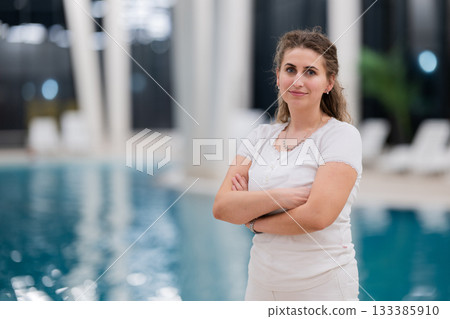 Portrait of a young female wellness coach smiling confidently with arms crossed at an indoor swimming pool, symbolizing health, fitness, and professional guidance. 133385910