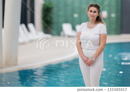 Portrait of a young female wellness coach smiling confidently at an indoor swimming pool, symbolizing health, fitness, and professional guidance. Portrait of a young female wellness coach smiling confidently at an indoor swimming pool, symbolizing health, fitness, and professional guidance. 133385917