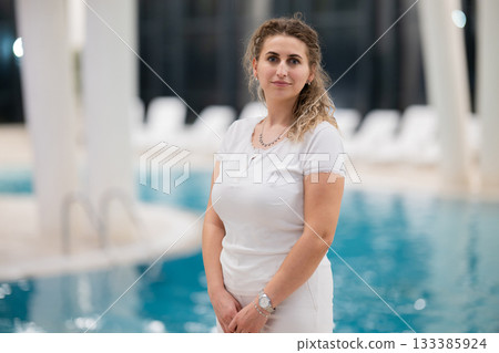 Portrait of a young female wellness coach smiling confidently at an indoor swimming pool, symbolizing health, fitness, and professional guidance. 133385924