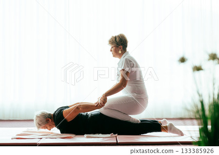Professional female physiotherapist supporting an older woman during a therapeutic stretching exercise on a mat. Rehabilitation and physiotherapy concept focused on mobility, flexibility, recovery Professional female physiotherapist supporting an older woman during a therapeutic stretching exercise on a mat. Rehabilitation and physiotherapy concept focused on mobility, flexibility, recovery 133385926