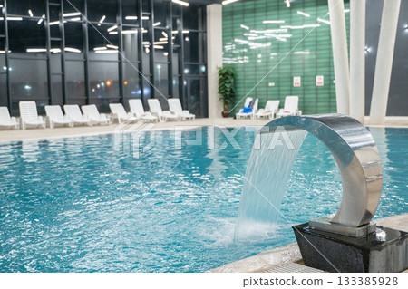 Contemporary indoor swimming pool with clean blue water, stylish lighting, and a metal waterfall fountain surrounded by white lounge chairs. Concept of wellness, luxury, relaxation, and modern spa 133385928