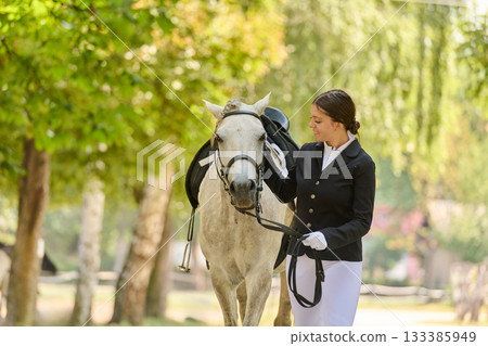 Beautiful young woman in riding clothes walking with her white horse in a park, equestrian lifestyle, sunny day, outdoor activity, nature background. Beautiful young woman in riding clothes walking with her white horse in a park, equestrian lifestyle, sunny day, outdoor activity, nature background. 133385949