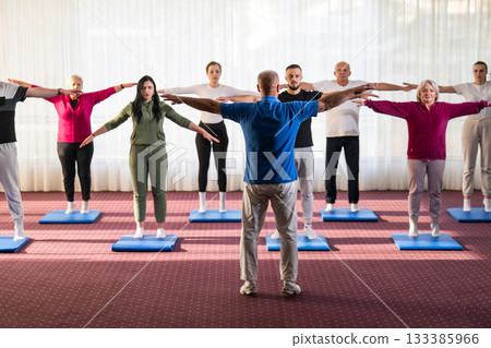 Instructor leading a diverse group of people during a fitness training session with stretching exercises on mats in a bright wellness center 133385966