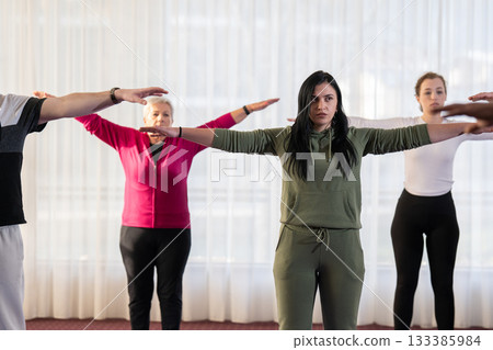 Group of people of different ages participating in a guided fitness or rehabilitation session indoors, standing on yoga mats with hands behind heads. 133385984
