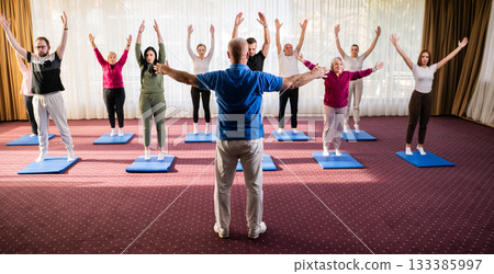 Instructor leading a diverse group of people during a fitness training session with stretching exercises on mats in a bright wellness center 133385997
