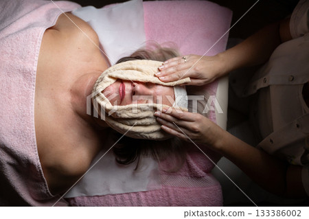 Close up of a mature woman receiving a relaxing facial massage from a professional female therapist in a modern spa. The client lies on a treatment bed with a towel and headband, enjoying skincare Close up of a mature woman receiving a relaxing facial massage from a professional female therapist in a modern spa. The client lies on a treatment bed with a towel and headband, enjoying skincare 133386002