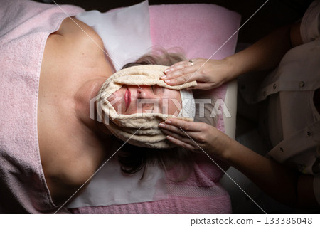 Close up of a mature woman receiving a relaxing facial massage from a professional female therapist in a modern spa. The client lies on a treatment bed with a towel and headband, enjoying skincare Close up of a mature woman receiving a relaxing facial massage from a professional female therapist in a modern spa. The client lies on a treatment bed with a towel and headband, enjoying skincare 133386048