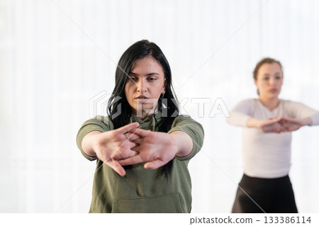 Focused young woman in a white shirt performing a stretching exercise indoors. Concept of wellness, health, flexibility, mindfulness, and active lifestyle. Focused young woman in a white shirt performing a stretching exercise indoors. Concept of wellness, health, flexibility, mindfulness, and active lifestyle. 133386114