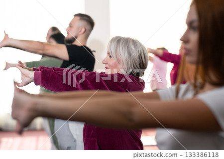 Group of people of different ages participating in a guided fitness or rehabilitation session indoors, standing on yoga mats with hands behind heads. Group of people of different ages participating in a guided fitness or rehabilitation session indoors, standing on yoga mats with hands behind heads. 133386118