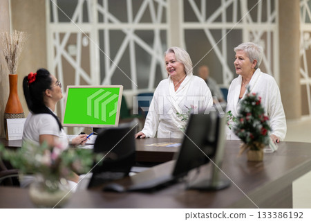 Two elderly women in white bathrobes smiling and talking to a receptionist at a wellness and spa center, representing hospitality, relaxation, and healthy lifestyle. Two elderly women in white bathrobes smiling and talking to a receptionist at a wellness and spa center, representing hospitality, relaxation, and healthy lifestyle. 133386192