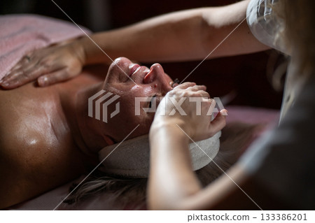 Close up of a mature woman receiving a relaxing facial massage from a professional female therapist in a modern spa. The client lies on a treatment bed with a towel and headband, enjoying skincare Close up of a mature woman receiving a relaxing facial massage from a professional female therapist in a modern spa. The client lies on a treatment bed with a towel and headband, enjoying skincare 133386201