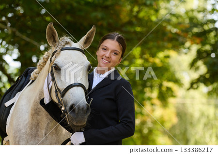 Smiling equestrian woman with her white horse in a natural outdoor setting, horse riding and equestrian sports, beautiful sunlight and green background. Smiling equestrian woman with her white horse in a natural outdoor setting, horse riding and equestrian sports, beautiful sunlight and green background. 133386217