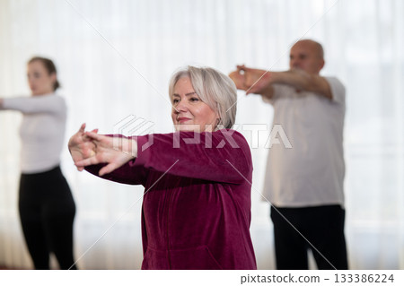 Active elderly woman in sportswear stretching her arms forward during a group fitness or rehabilitation session. Concept of healthy aging, wellness, flexibility, and active senior lifestyle. Active elderly woman in sportswear stretching her arms forward during a group fitness or rehabilitation session. Concept of healthy aging, wellness, flexibility, and active senior lifestyle. 133386224