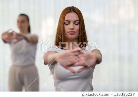 Focused young woman in a white shirt performing a stretching exercise indoors. Concept of wellness, health, flexibility, mindfulness, and active lifestyle. Focused young woman in a white shirt performing a stretching exercise indoors. Concept of wellness, health, flexibility, mindfulness, and active lifestyle. 133386227
