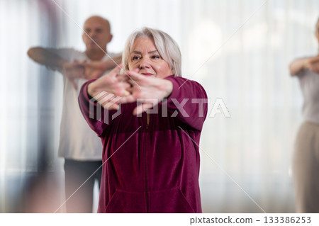 Active elderly woman in sportswear stretching her arms forward during a group fitness or rehabilitation session. Concept of healthy aging, wellness, flexibility, and active senior lifestyle. 133386253