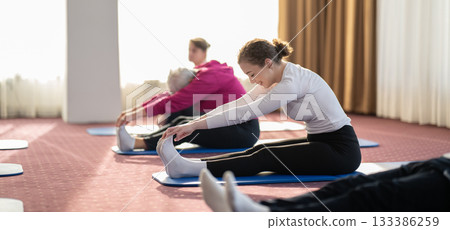 Women of different ages stretching forward on yoga mats during a group fitness or wellness session indoors. Concept of flexibility, mindfulness, physical health, and relaxation through stretching Women of different ages stretching forward on yoga mats during a group fitness or wellness session indoors. Concept of flexibility, mindfulness, physical health, and relaxation through stretching 133386259