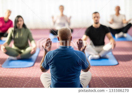 Rear view of a male instructor guiding a mixed age group during a stretching or rehabilitation class indoors. Participants are sitting on yoga mats following his movements. Concept of health, wellness Rear view of a male instructor guiding a mixed age group during a stretching or rehabilitation class indoors. Participants are sitting on yoga mats following his movements. Concept of health, wellness 133386270