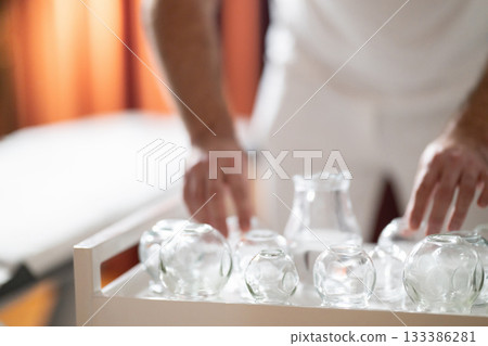 Therapist Preparing Glass Cups for Traditional Cupping Therapy. Close up of therapists hands arranging clean glass cups on a tray before starting a traditional cupping therapy session. Therapist Preparing Glass Cups for Traditional Cupping Therapy. Close up of therapists hands arranging clean glass cups on a tray before starting a traditional cupping therapy session. 133386281