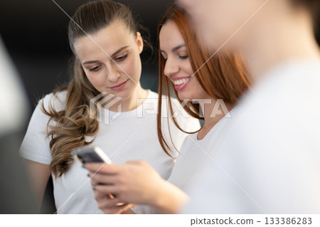 Two young women in casual white shirts smiling and looking at a smartphone screen together indoors. Concept of friendship, social connection, communication, and modern lifestyle. 133386283