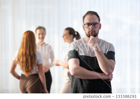 Portrait of a confident young man wearing glasses and smiling while standing with his colleagues in the background during an informal business meeting. Portrait of a confident young man wearing glasses and smiling while standing with his colleagues in the background during an informal business meeting. 133386291