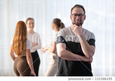 Portrait of a confident young man wearing glasses and smiling while standing with his colleagues in the background during an informal business meeting. 133386295