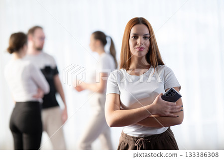 Confident young woman holding a smartphone while standing in front of colleagues having a discussion in the background. Concept of leadership, communication, and modern teamwork. Confident young woman holding a smartphone while standing in front of colleagues having a discussion in the background. Concept of leadership, communication, and modern teamwork. 133386308