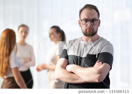 Portrait of a confident young man with glasses smiling and standing with folded arms while his colleagues talk in the background. Concept of leadership, teamwork, and success in a modern professional Portrait of a confident young man with glasses smiling and standing with folded arms while his colleagues talk in the background. Concept of leadership, teamwork, and success in a modern professional 133386313