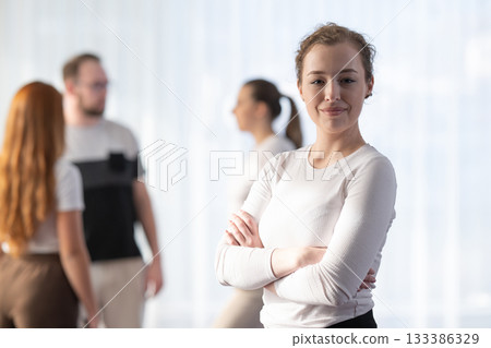 Portrait of a confident woman standing with arms crossed and smiling at the camera while her colleagues interact in the blurred background. Portrait of a confident woman standing with arms crossed and smiling at the camera while her colleagues interact in the blurred background. 133386329