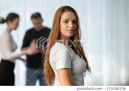 Portrait of a confident young woman standing in a bright modern workspace with colleagues conversing in the background. Concept of leadership, independence, and professional success. Portrait of a confident young woman standing in a bright modern workspace with colleagues conversing in the background. Concept of leadership, independence, and professional success. 133386336