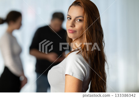 Portrait of a confident young woman with long hair looking at the camera, while people talk in the blurred background. Concept of confidence, individuality, beauty, focus, and empowerment. Portrait of a confident young woman with long hair looking at the camera, while people talk in the blurred background. Concept of confidence, individuality, beauty, focus, and empowerment. 133386342