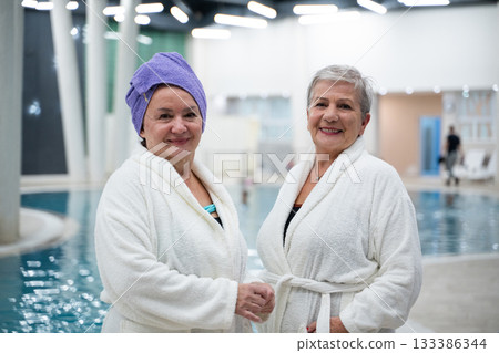 Two cheerful senior women in white bathrobes laughing and relaxing together at an indoor swimming pool. Concept of friendship, wellness, spa lifestyle, and healthy aging. Two cheerful senior women in white bathrobes laughing and relaxing together at an indoor swimming pool. Concept of friendship, wellness, spa lifestyle, and healthy aging. 133386344