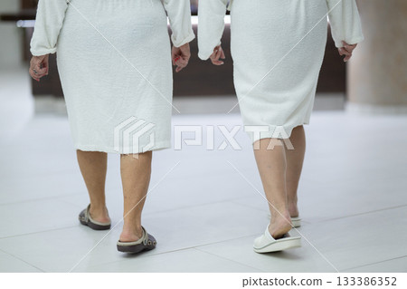 Elderly women walking together in a wellness and spa center dressed in white bathrobes, symbolizing relaxation, friendship, and healthy lifestyle. 133386352