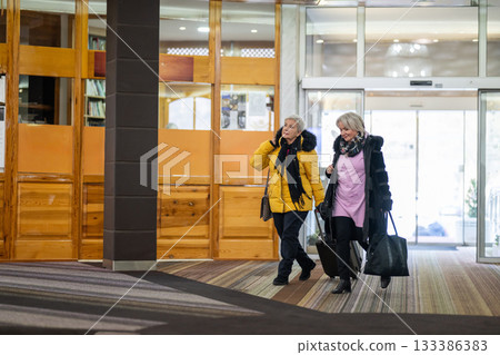 Two older women dressed in winter clothing standing in a hotel lobby and having a friendly conversation next to their luggage. Concept of travel, friendship, and leisure lifestyle for seniors. 133386383